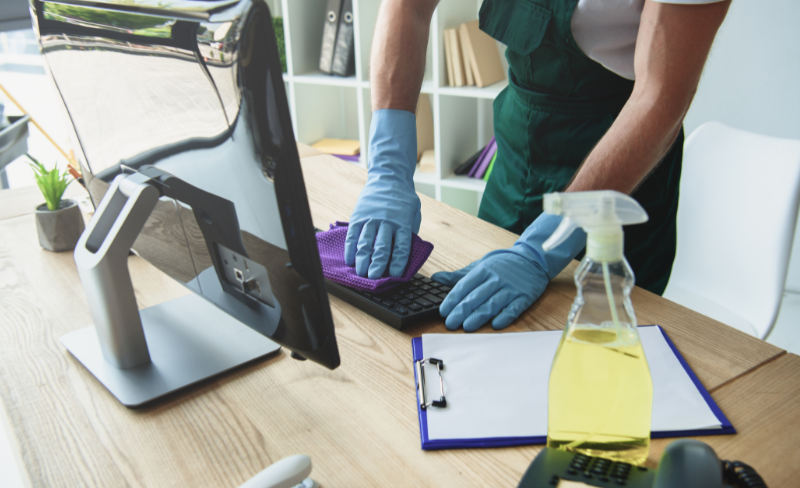 Technician sanitizing high-touch surfaces including desks, keyboards, and door handles.